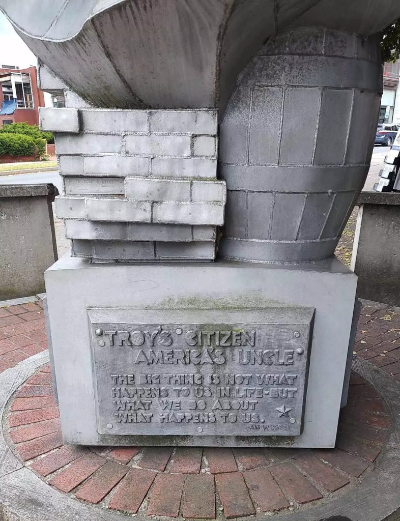 Base of Uncle Sam statue in Troy, New York, featuring a relief of bricks, a barrel, and an engraved plaque reading “Troy's Citizen, America's Uncle…”